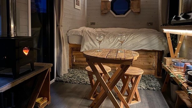 A cozy room with a bed, a table with two chairs, a fireplace, and a window with a view of the night sky. - 1 Bed Cabin - Sleeps 2 - Hottub - Parking - Garden (Bideford)