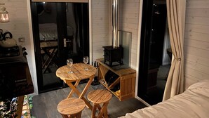 A cozy room with a wooden interior, a small table with two stools, a bed, and a wood-burning stove. - 1 Bed Cabin - Sleeps 2 - Hottub - Parking - Garden (Bideford)