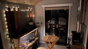 A cozy room with a wooden ceiling, a kitchen area with a stove, a dining table with chairs, and a large window. - 1 Bed Cabin - Sleeps 2 - Hottub - Parking - Garden (Bideford)