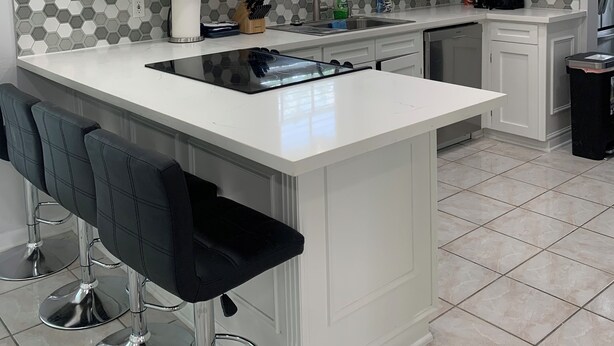 Une cuisine moderne avec des meubles blancs, un grand îlot central doté de tabourets, une cuisinière encastrée et une fenêtre avec des stores.