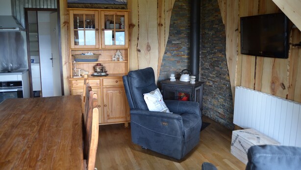 A cozy living room with a wooden dining table, a black leather sofa, a wooden cabinet with glass doors, a fireplace, and a television mounted on the wall. - Haut de Chalet Le Bois de Cerf , Animaux Acceptés en Lisière de Foret (Les Angles)