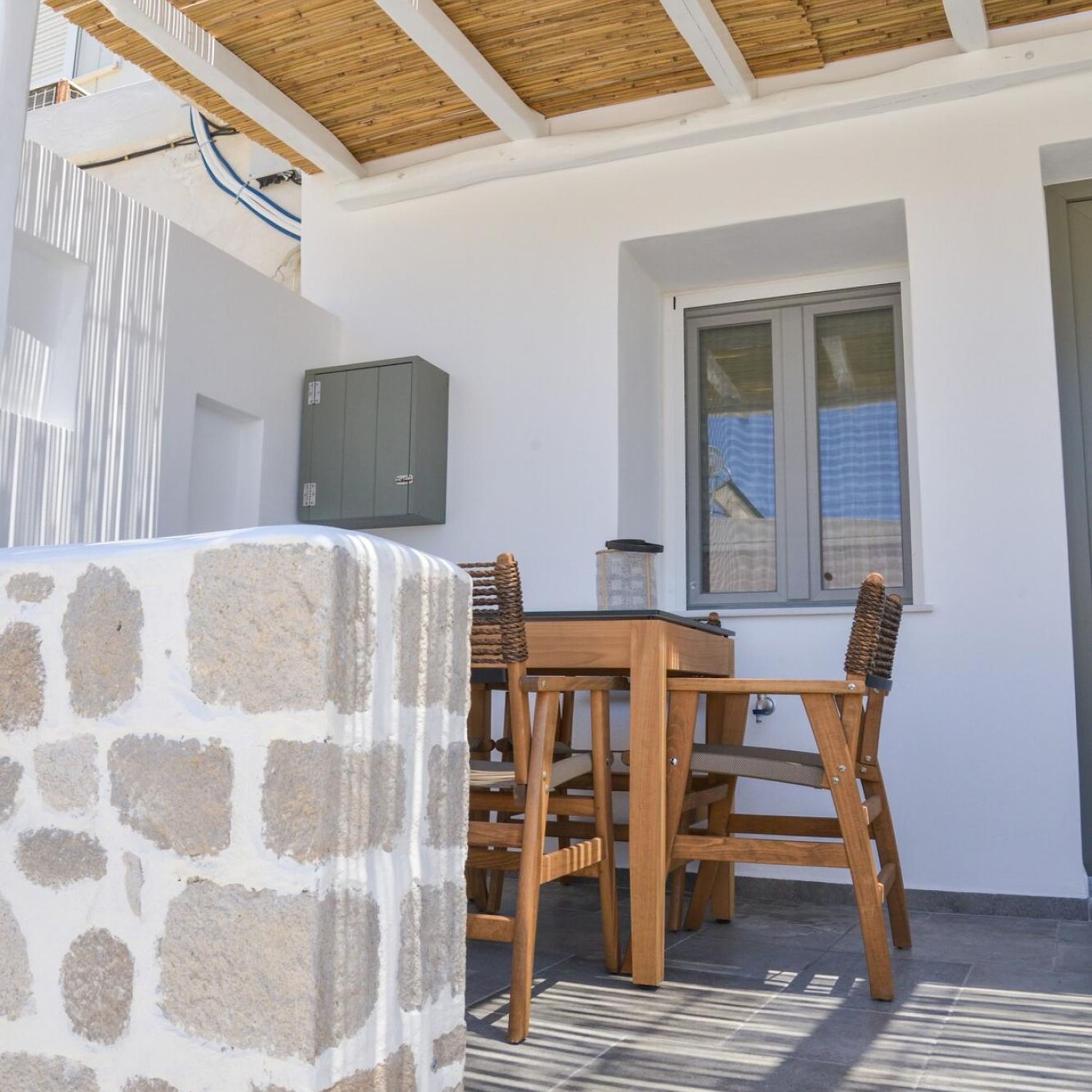 Pared de piedra, mesa de madera y sillas en un espacio al aire libre cubierto, con una ventana y un cuadro eléctrico fijado a la pared.