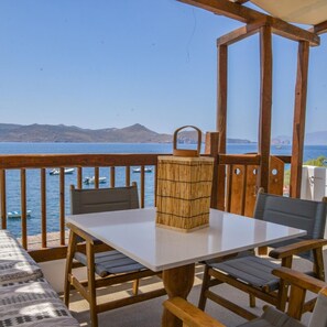 A wooden deck with a table and chairs, overlooking a body of water with boats and distant mountains. - Honeymoon Klima Suite (Milos)