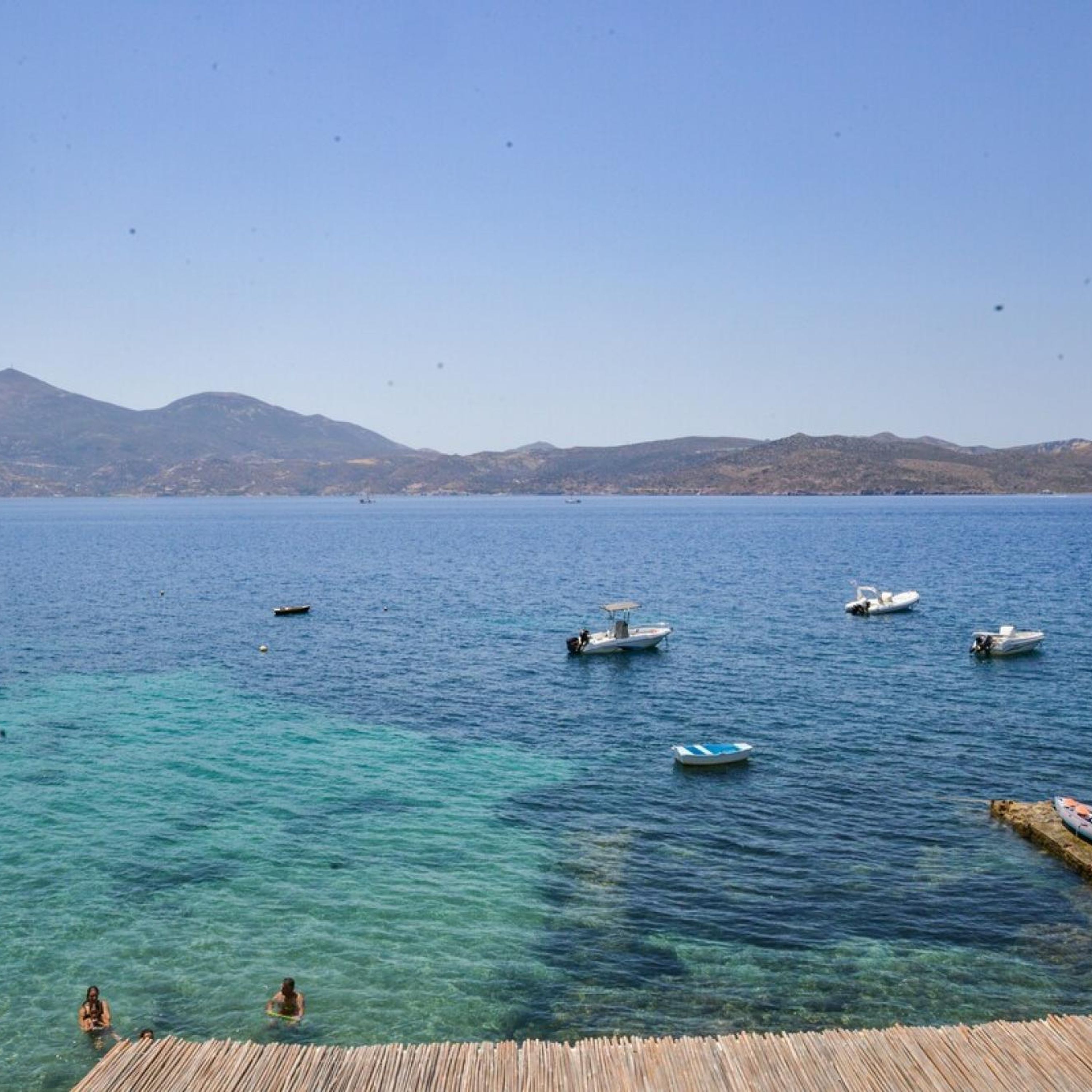 A calm sea with several boats, a wooden dock, and a mountainous backdrop.