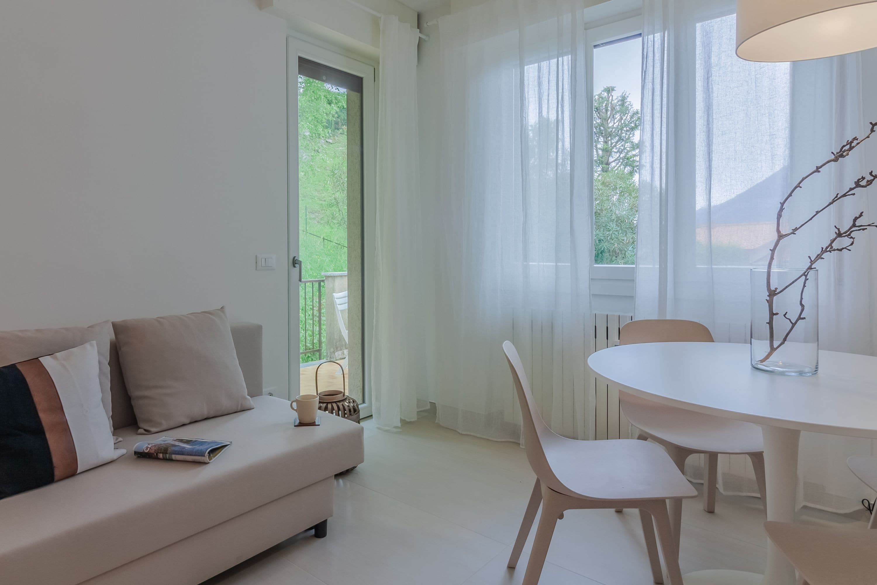 A modern living room with a sofa, a dining table, chairs, and a view of greenery through the windows.