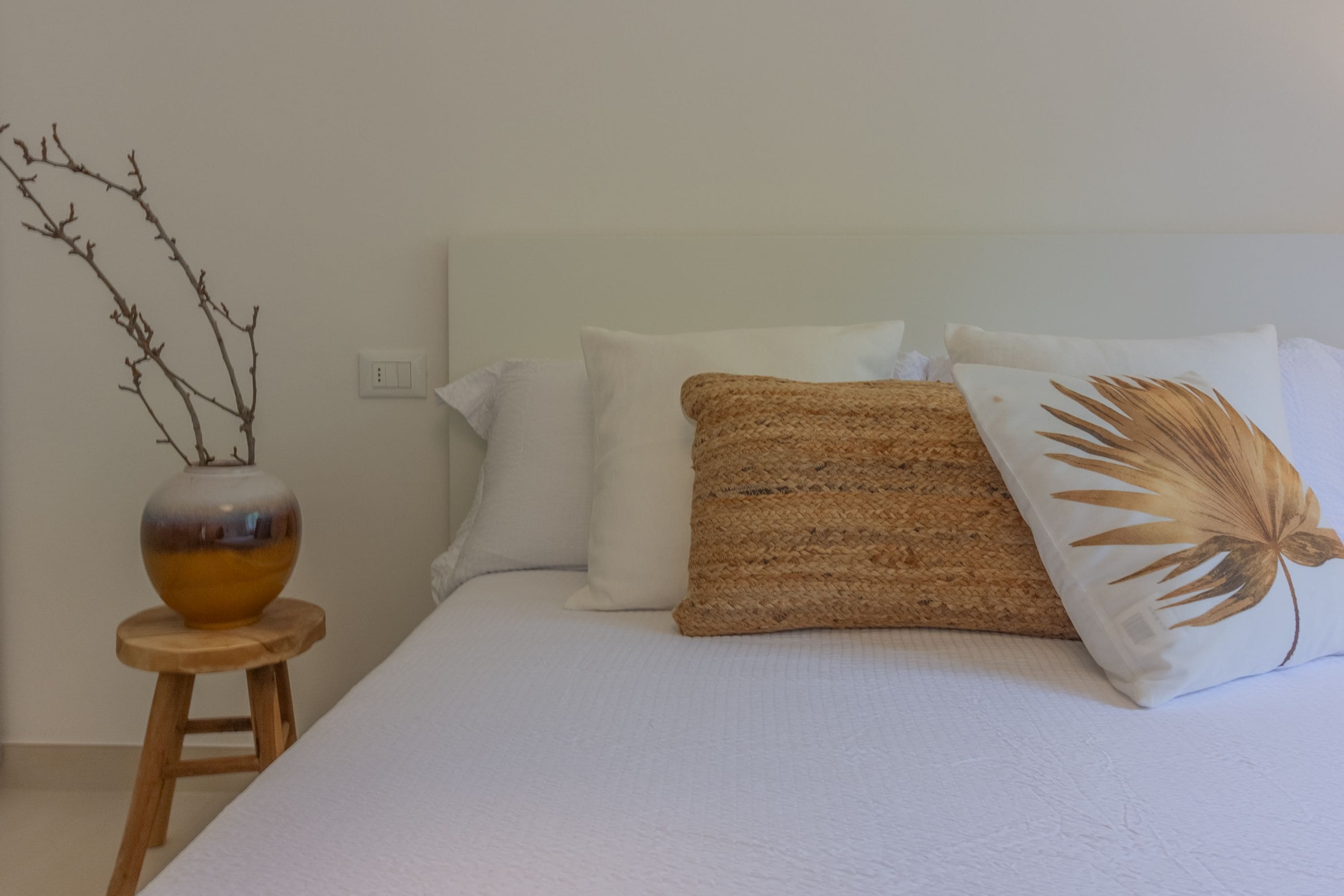 A bed with white bedding, a wooden stool, and a vase with branches.