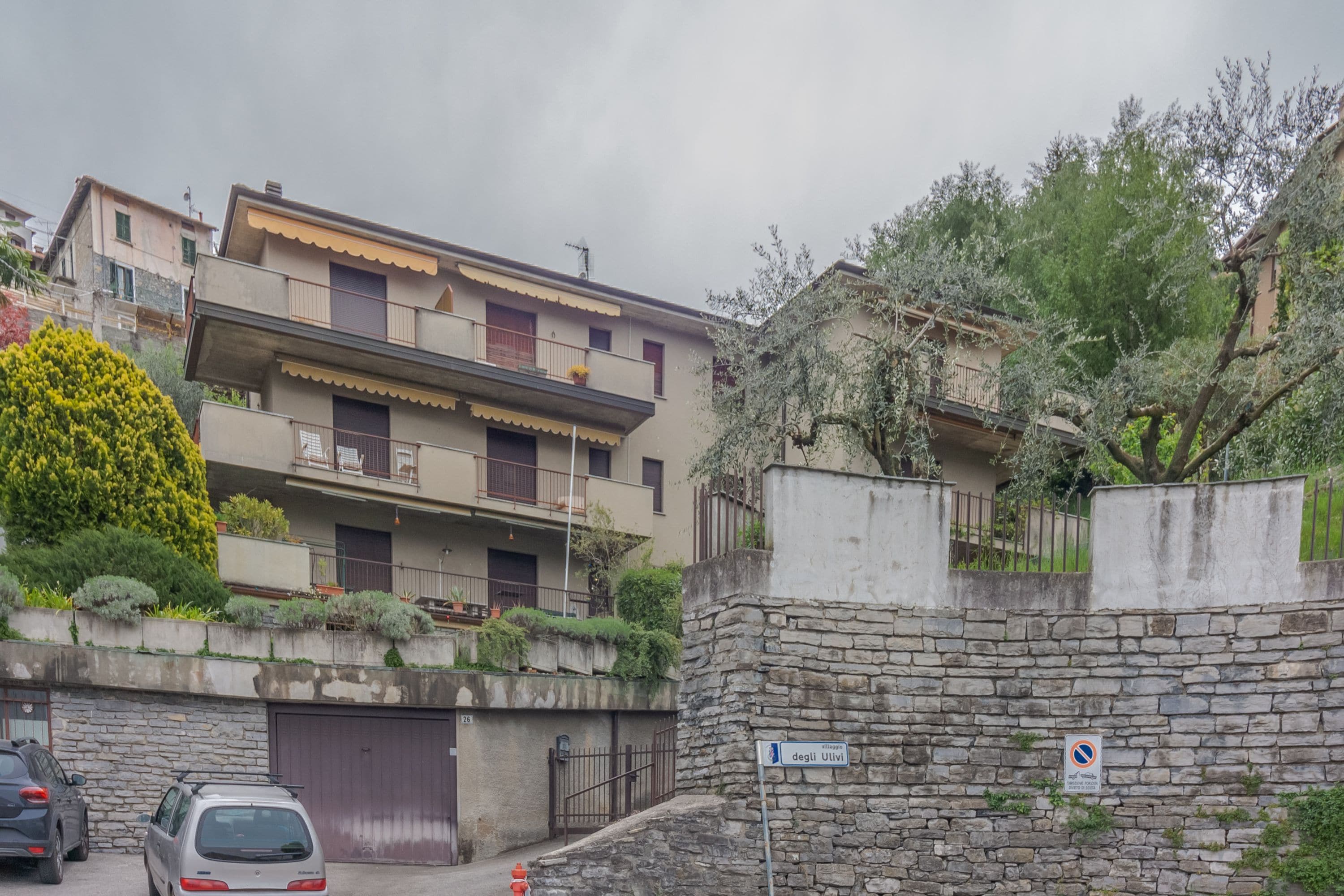 A multi-story residential building with balconies, a stone wall, and a parking area.