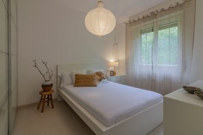 Un dormitorio con cama, mesita de noche, lámpara, ventana y un jarrón decorativo.