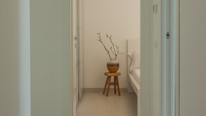 Un couloir avec une porte blanche, un petit tabouret sur lequel est posé un vase, et un lit à l’arrière-plan.