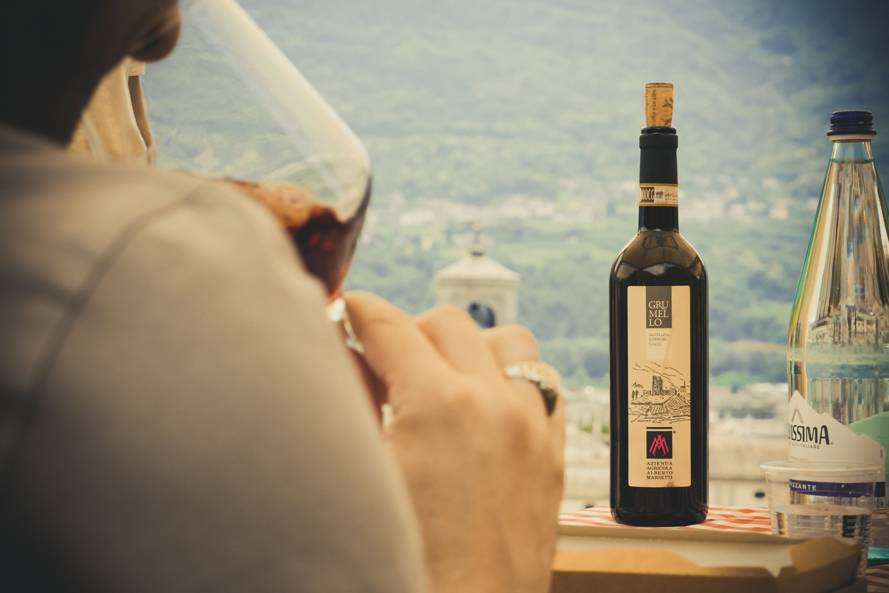 A person holding a wine glass, a bottle of Grumelo wine, and a bottle of Missima water on a table with a scenic view in the background.