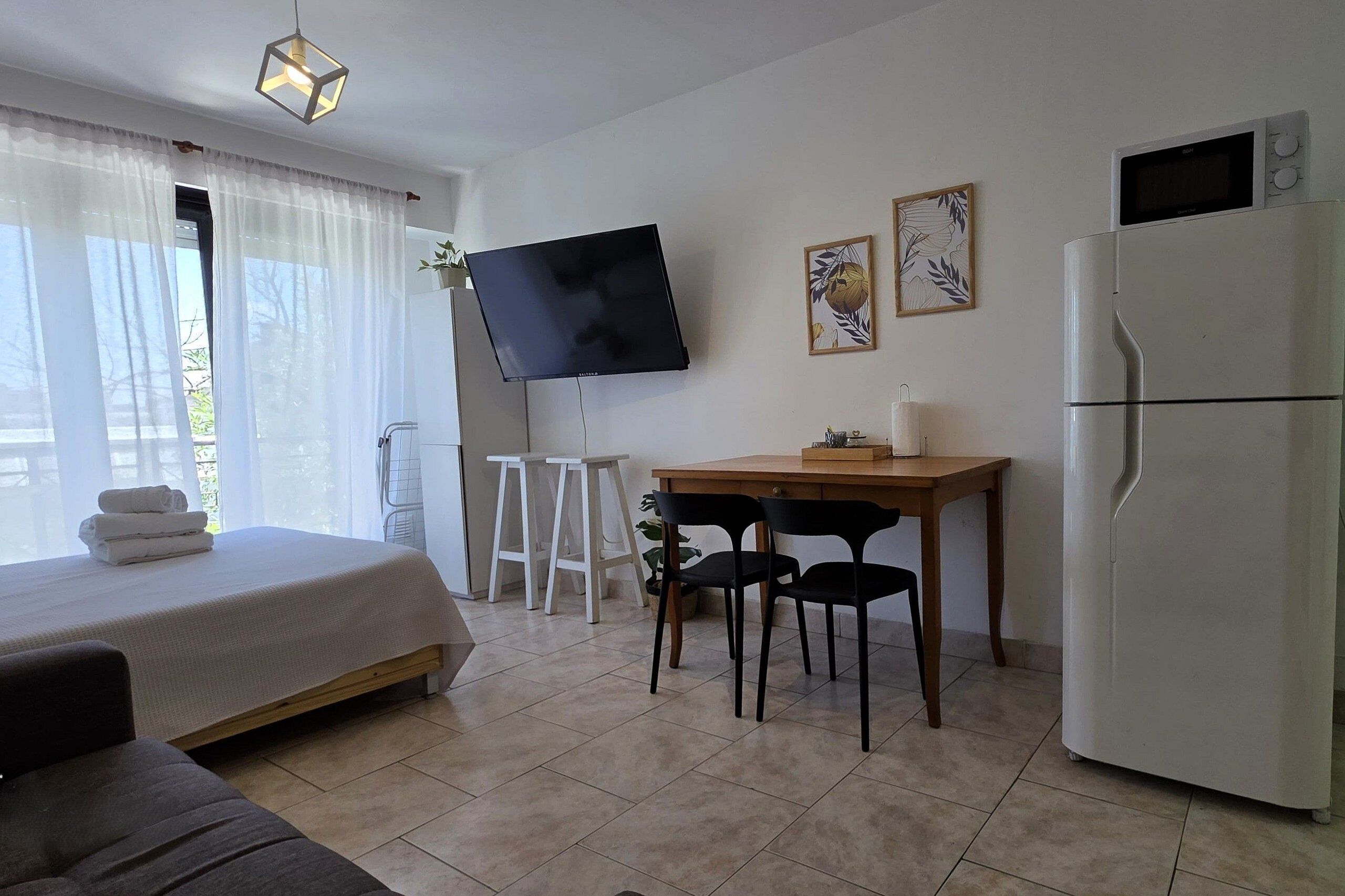 A hotel room with a bed, a desk with two chairs, a flat-screen TV, a refrigerator, and a window with sheer curtains.