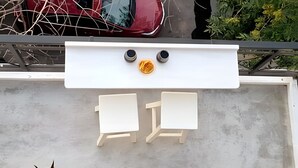 Een terras op het dak met twee witte stoelen en een kleine tafel, omgeven door potplanten en een rode auto op de achtergrond.