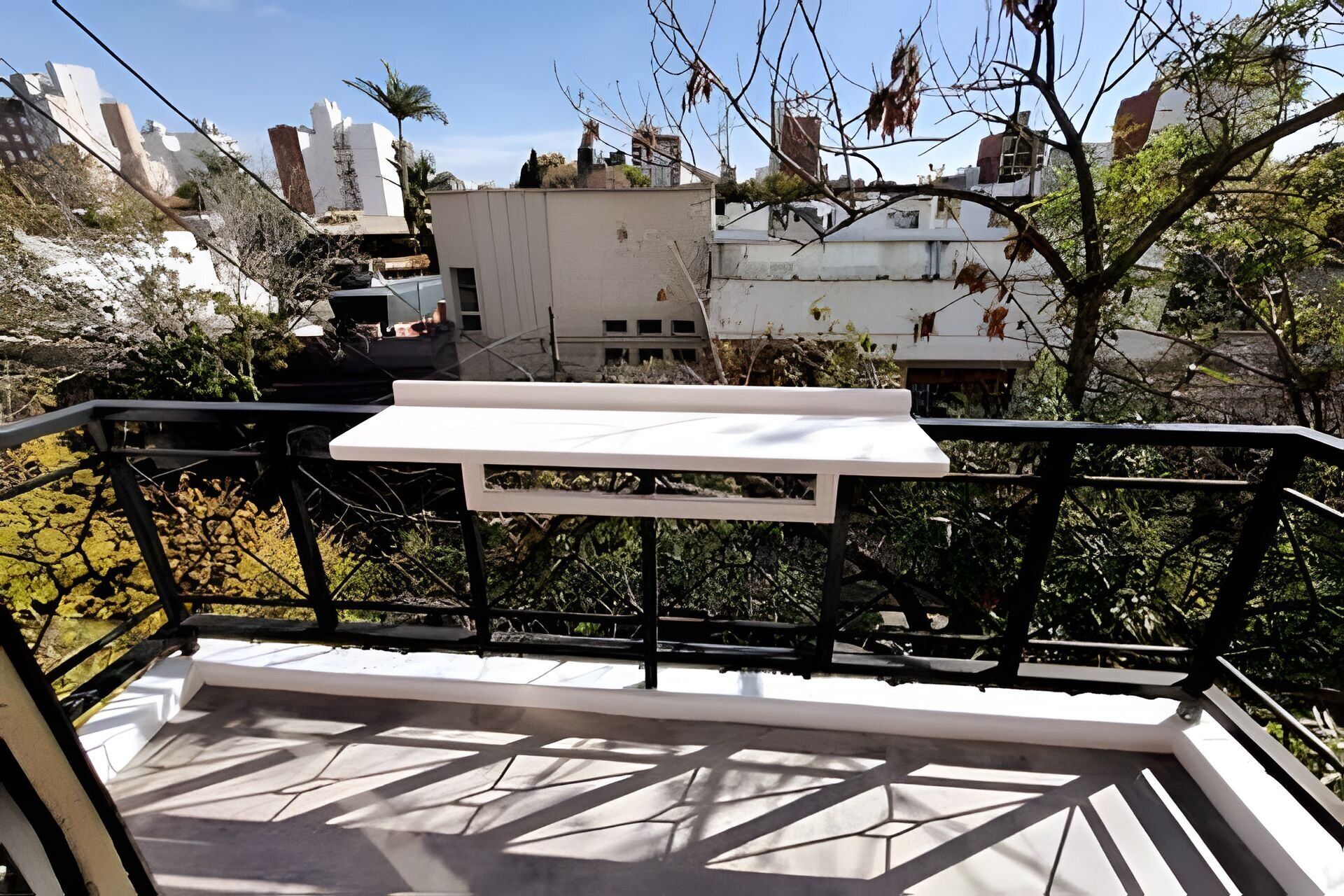 A balcony with a white table and black railing, overlooking a cityscape with buildings and trees.