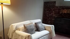 A white sofa with grey cushions in a room with a tiled floor, a standing floor lamp, and a stone wall. - Palace of Alboin (Pavia)