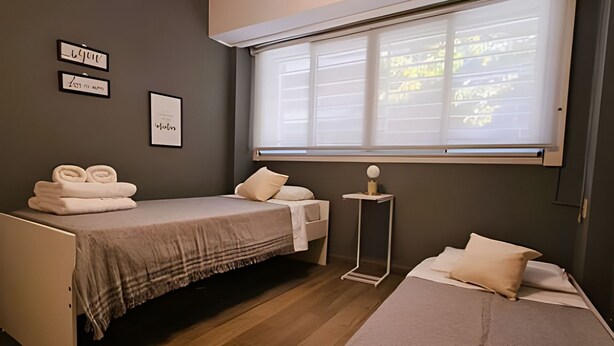 A bedroom with two beds, a small table, and a window with blinds. - Hornera Roca - Amplio y Cómodo, Pleno Microcentro (Rosario)