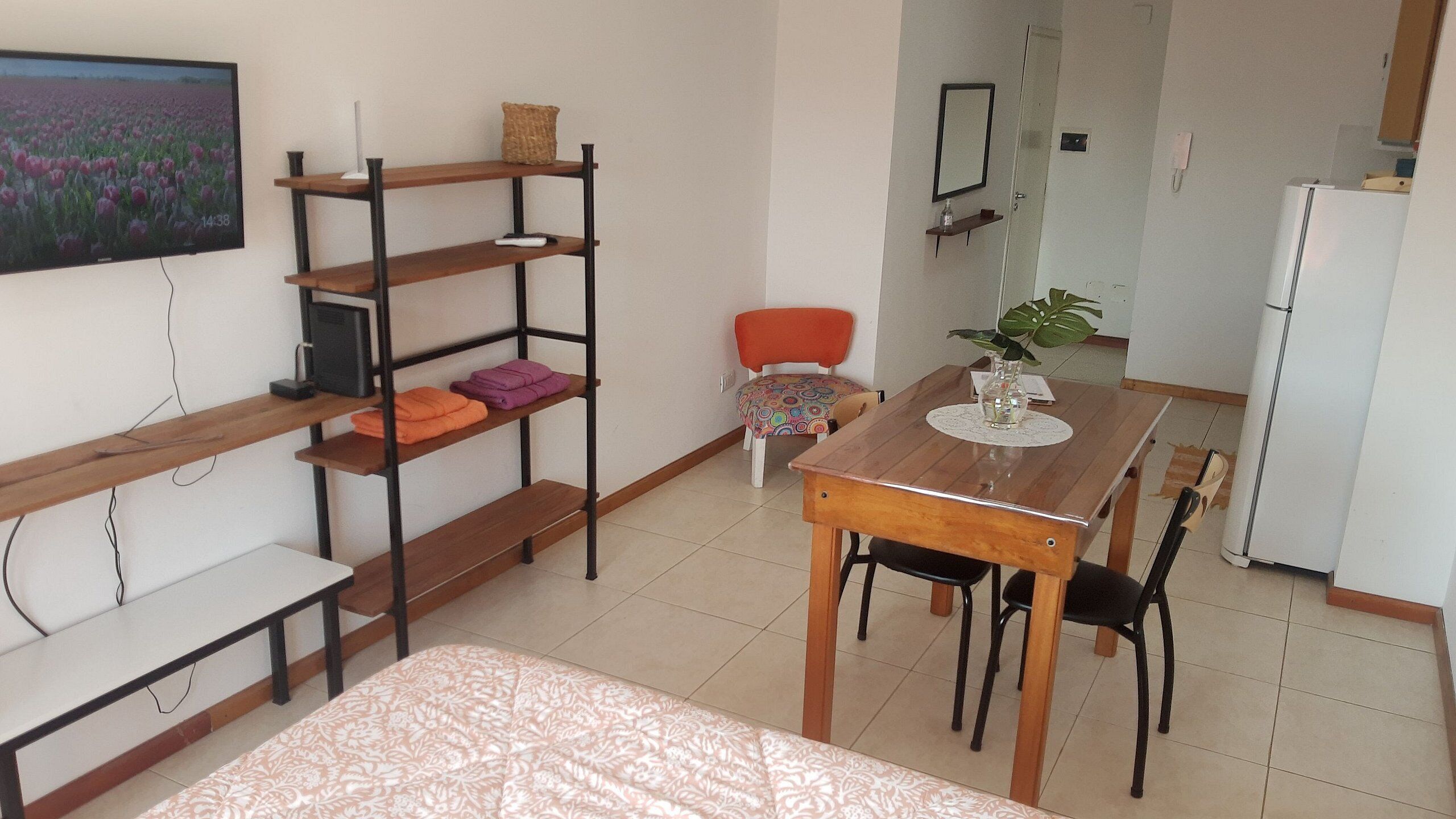 Quarto compacto com cama, mesa de madeira com cadeiras, TV em uma prateleira, poltrona com almofada colorida e geladeira.