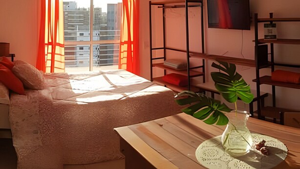A modern bedroom with a bed, a desk, a plant, and a view of buildings through the window. - Hornera Lagos - Modern, Spacious & Bright (Rosario)