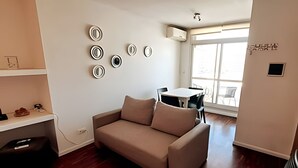 A modern living room with a beige sofa, a dining table with chairs, and a large window providing natural light. - Hornera San Juan - Bright Balcony Apartment (Rosario)
