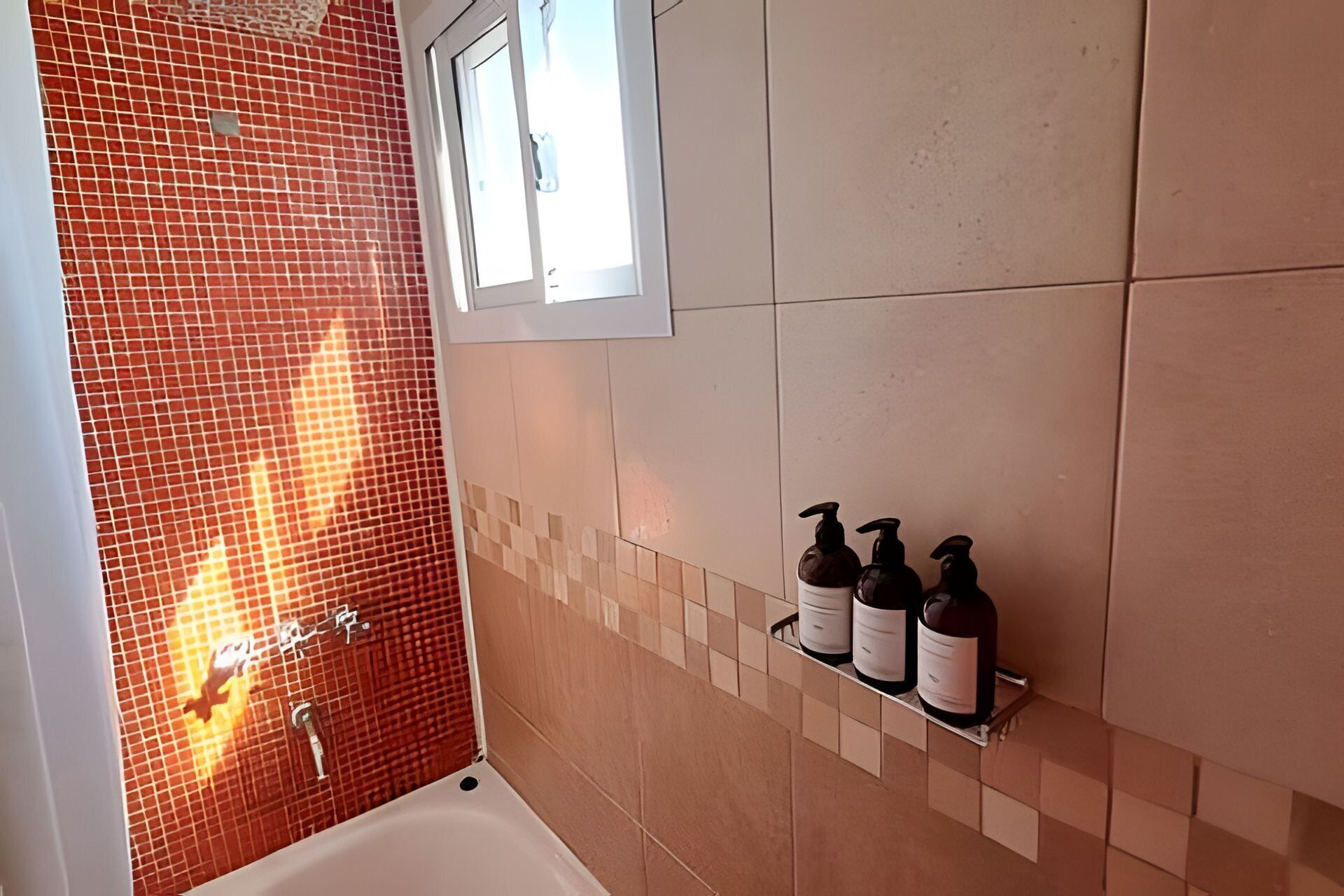 A bathroom with a red mosaic wall, a bathtub, and a shelf with three bottles.