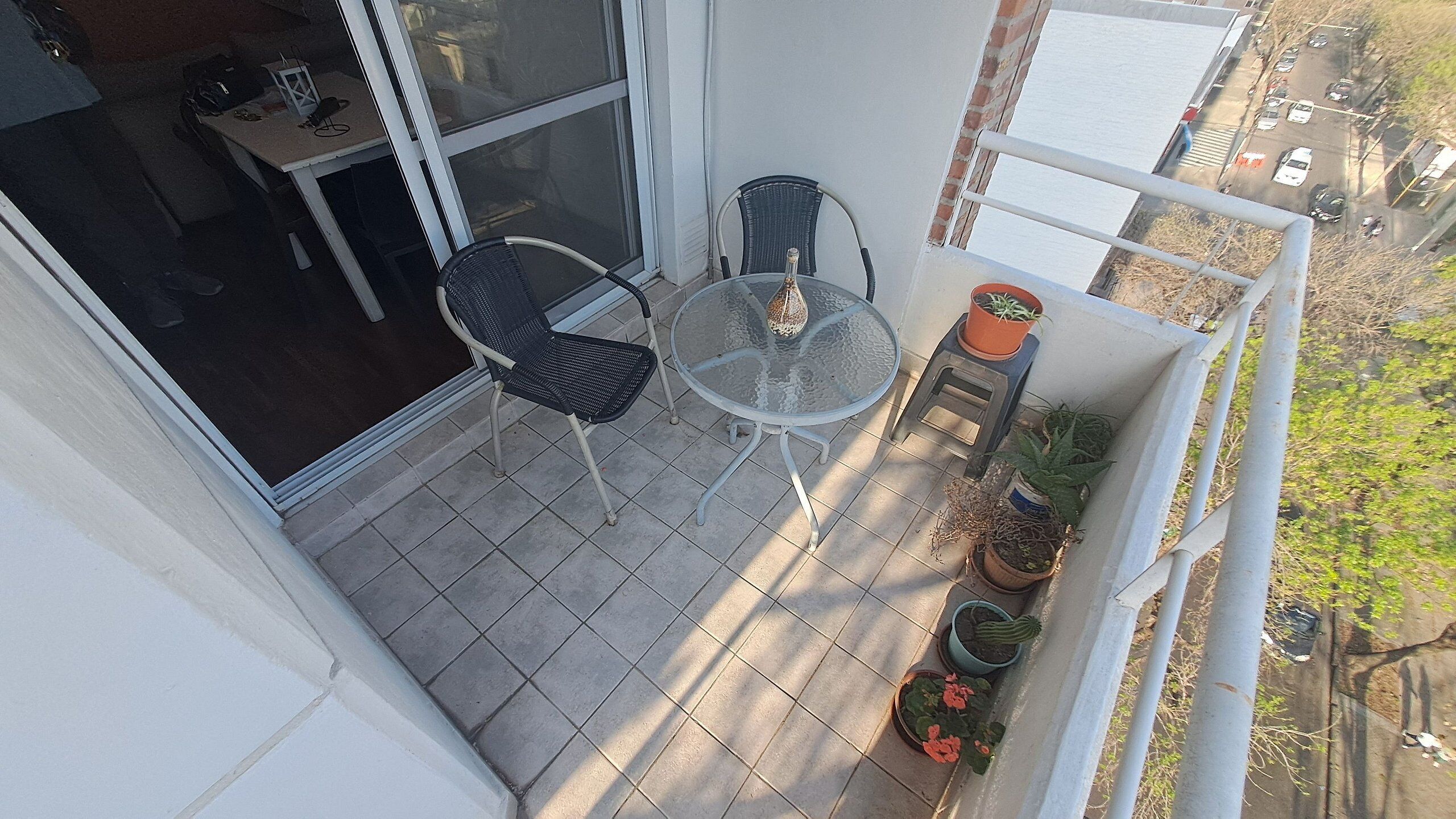 A balcony with a glass table and two chairs, potted plants, and a view of the street.