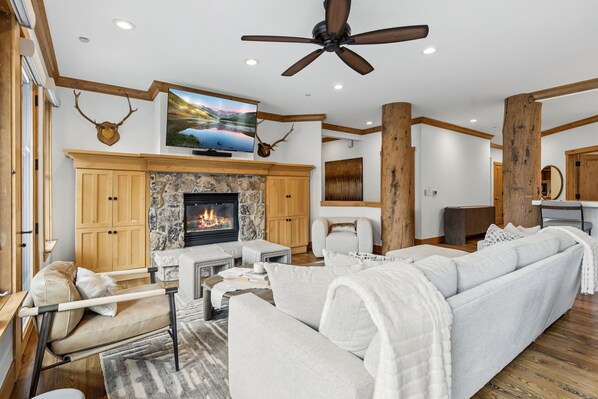 A spacious living room with a fireplace, a large sofa, a television, and a ceiling fan. - Modern Village Condo by InvitedHome | Pool, Hot Tub, Ski-in/Out, Breakfast (Beaver Creek)