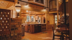 Accogliente interno di una baita in legno con zona cucina centrale, tavolo da pranzo e una scala che conduce a un piano superiore.
