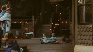 Eine Holzterrasse im Freien mit spielenden Kindern, einem Schild mit der Aufschrift 'TREEHOUSE PENTHAUS' und einer Ziegelwand.