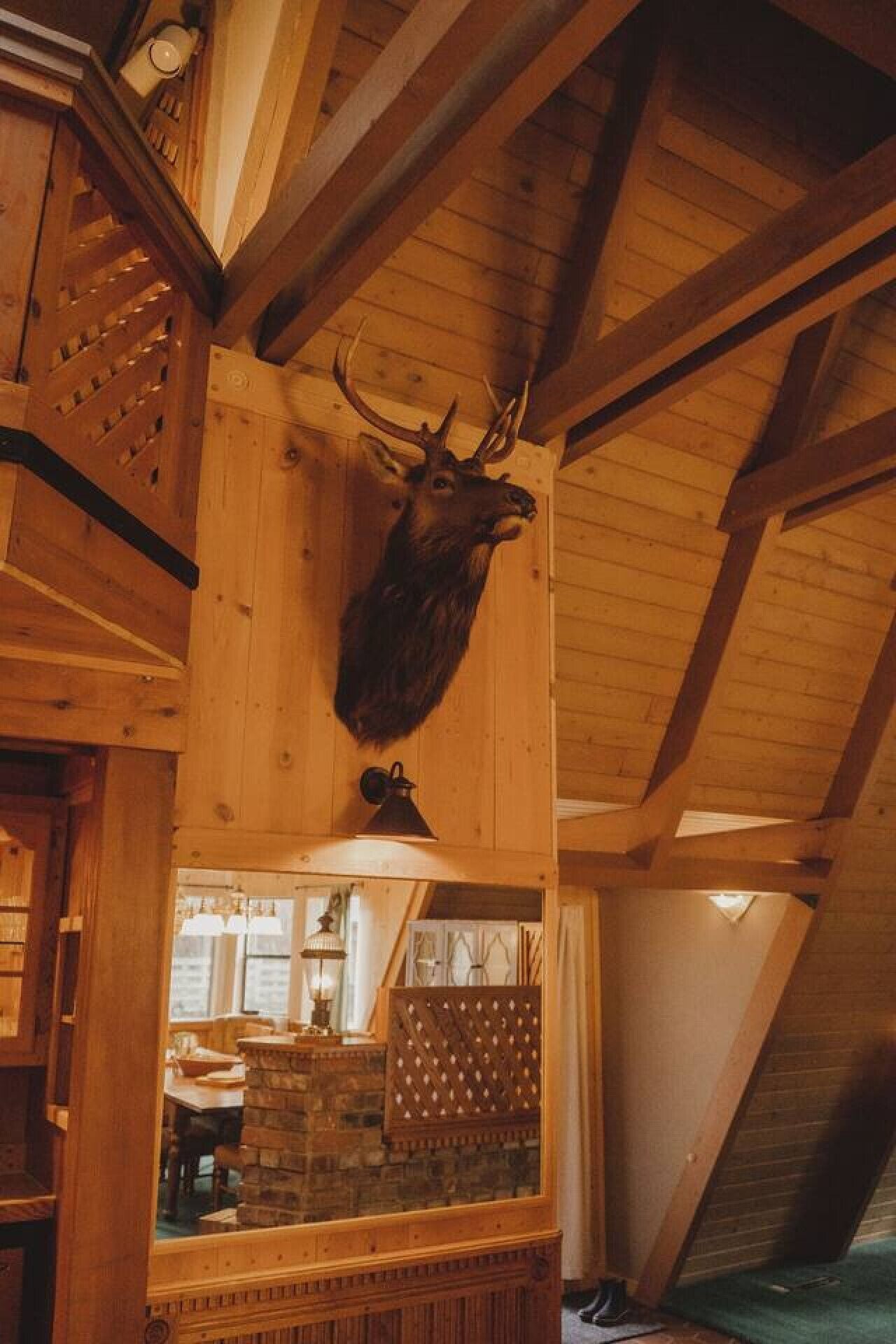 Un plafond en bois orné d’une tête de cerf fixée au mur, une cheminée en pierre et un coin repas visible à travers une fenêtre.
