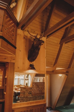 Interior de madera con un venado disecado en la pared y vigas de madera, junto a una chimenea de piedra.