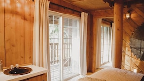 A wooden cabin interior with a large sliding glass door leading to a balcony, a white sink, and a cozy bed with a fluffy blanket.