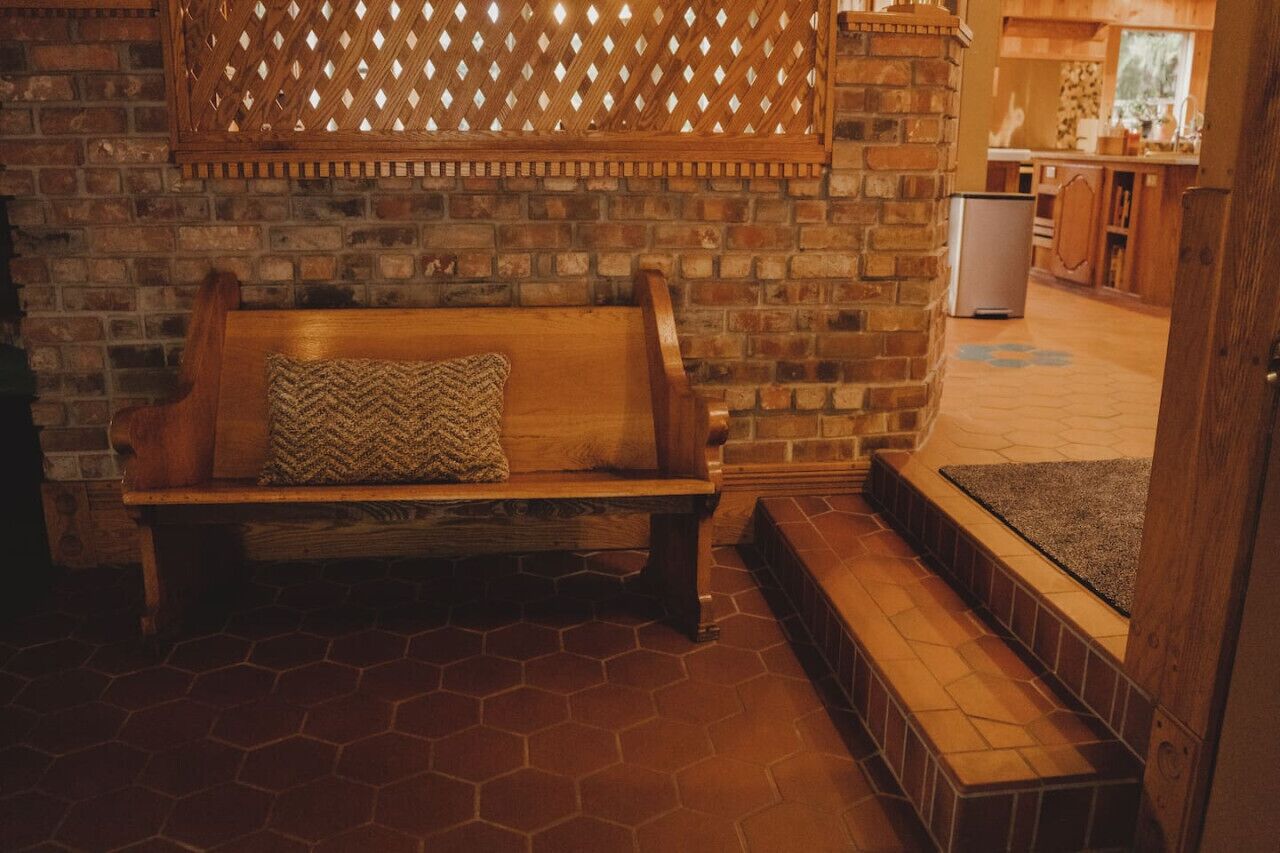 Un banc en bois avec un coussin tissé, dans une pièce dont le mur est en briques et le sol recouvert de carreaux hexagonaux.