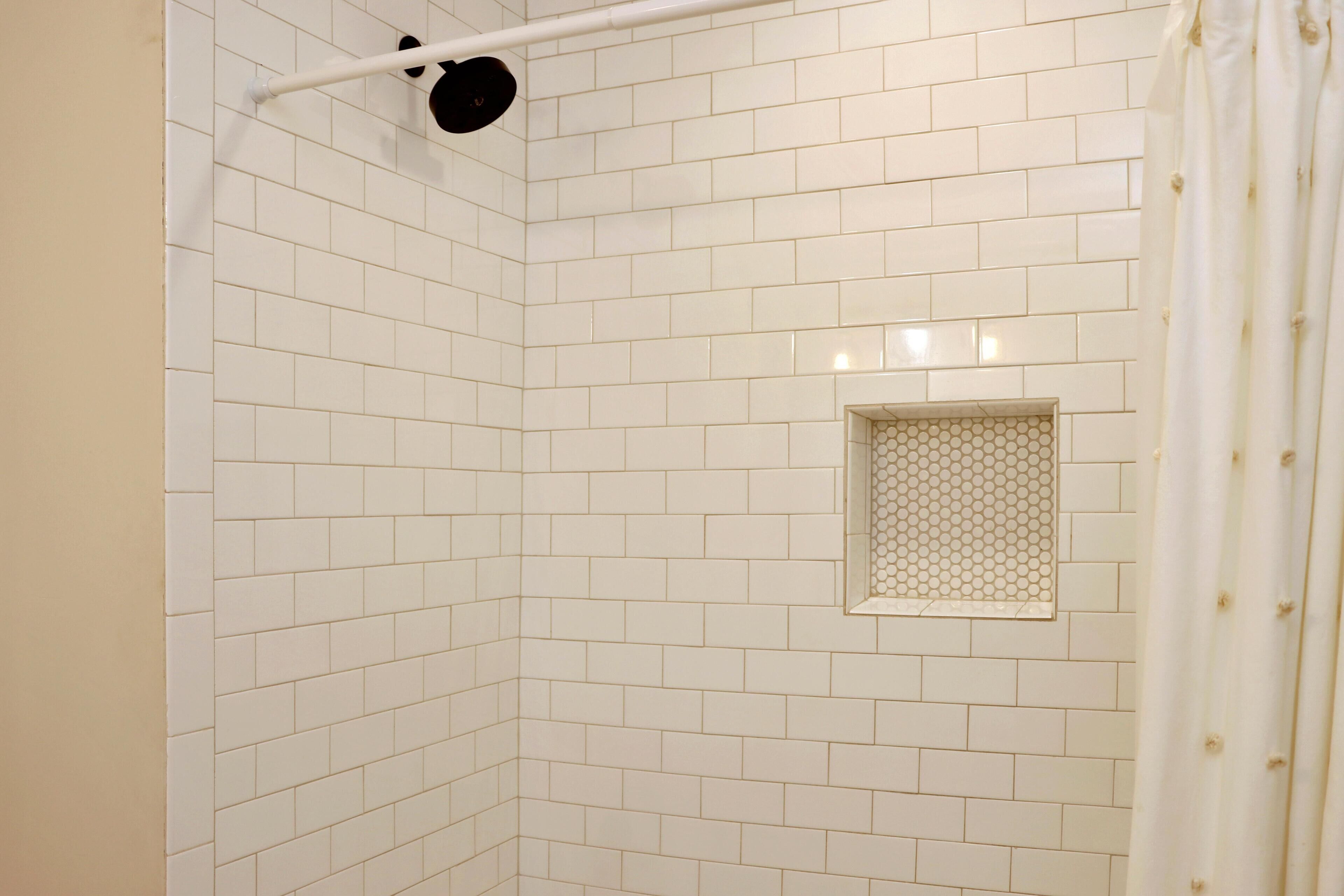 Un coin de salle de bain avec des carreaux de céramique blancs rectangulaires, un rideau de douche et une petite fenêtre ornée d’un motif en nid d’abeille.