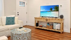 Un soggiorno con un divano bianco, un tavolino rotondo, un mobile TV in legno con una TV che mostra una spiaggia e una porta con una finestra.