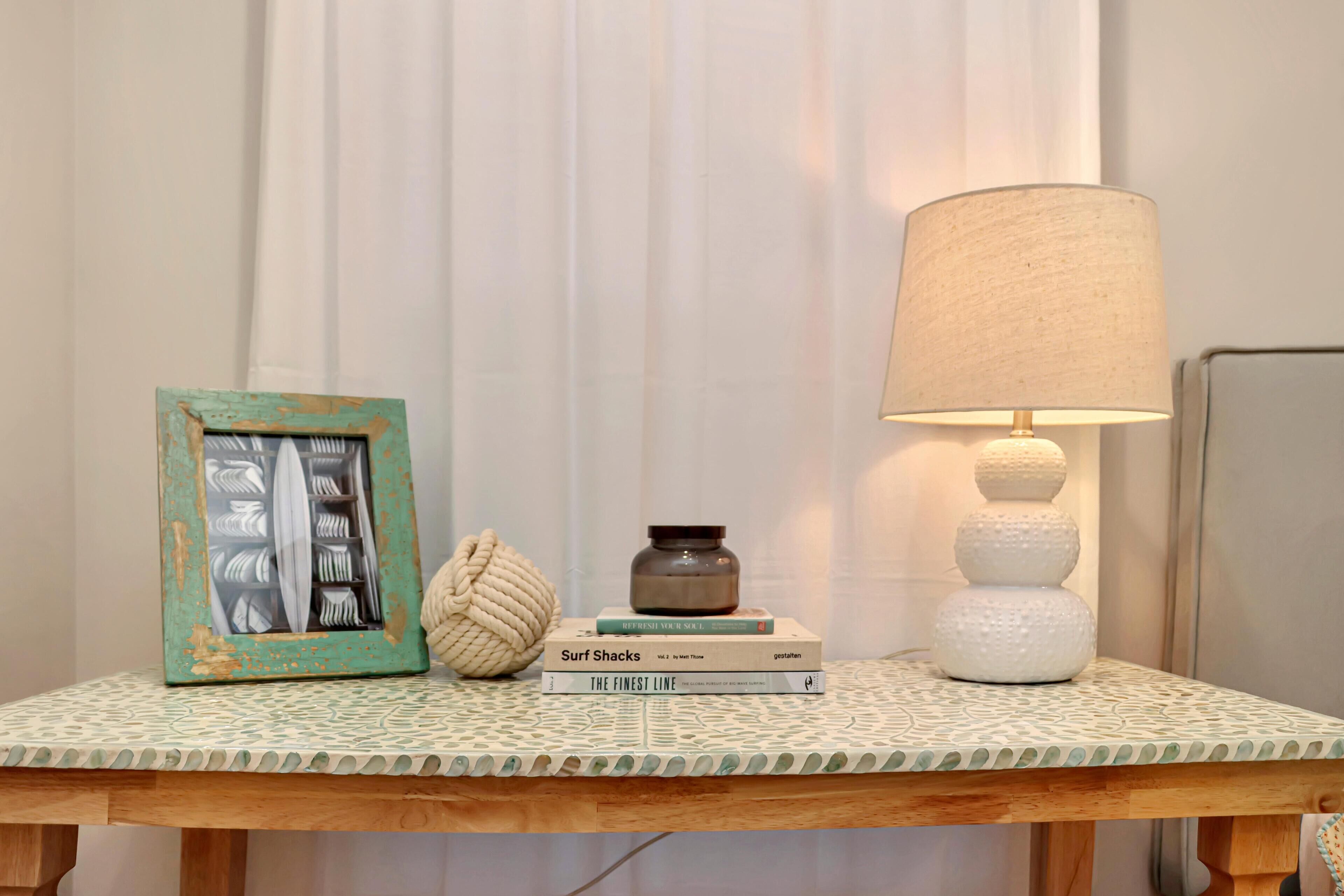 Een houten tafel met een tafelkleed met patroon, een lijst met afbeelding, een stapel boeken, een decoratieve lamp en een gevlekte vaas.