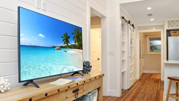 Un televisore su una consolle in legno con una spiaggia sullo schermo, interni bianchi e un cartello con su scritto 'Beach House'.