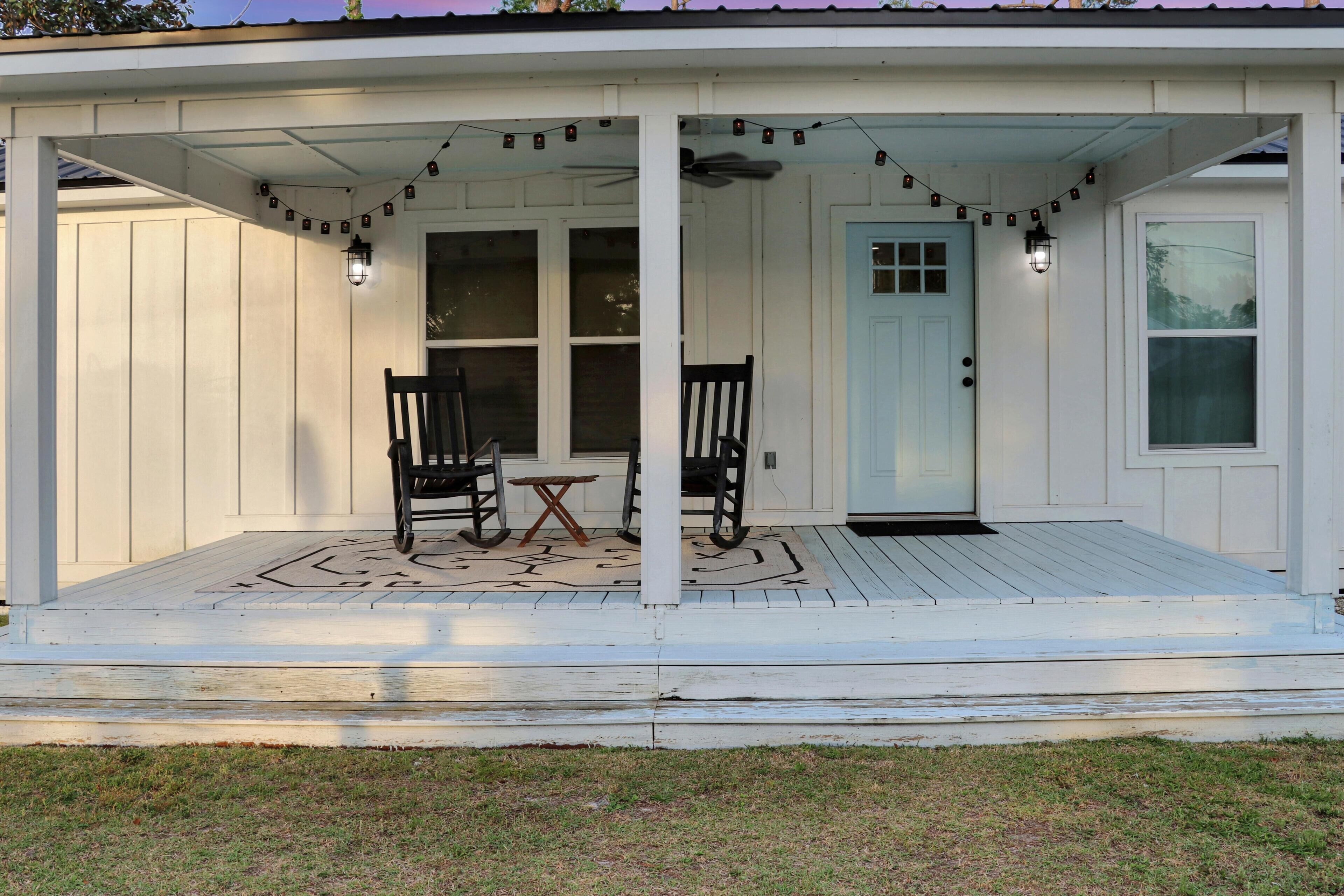 A covered porch with two rocking chairs and a small table, featuring string lights and a door with a window.