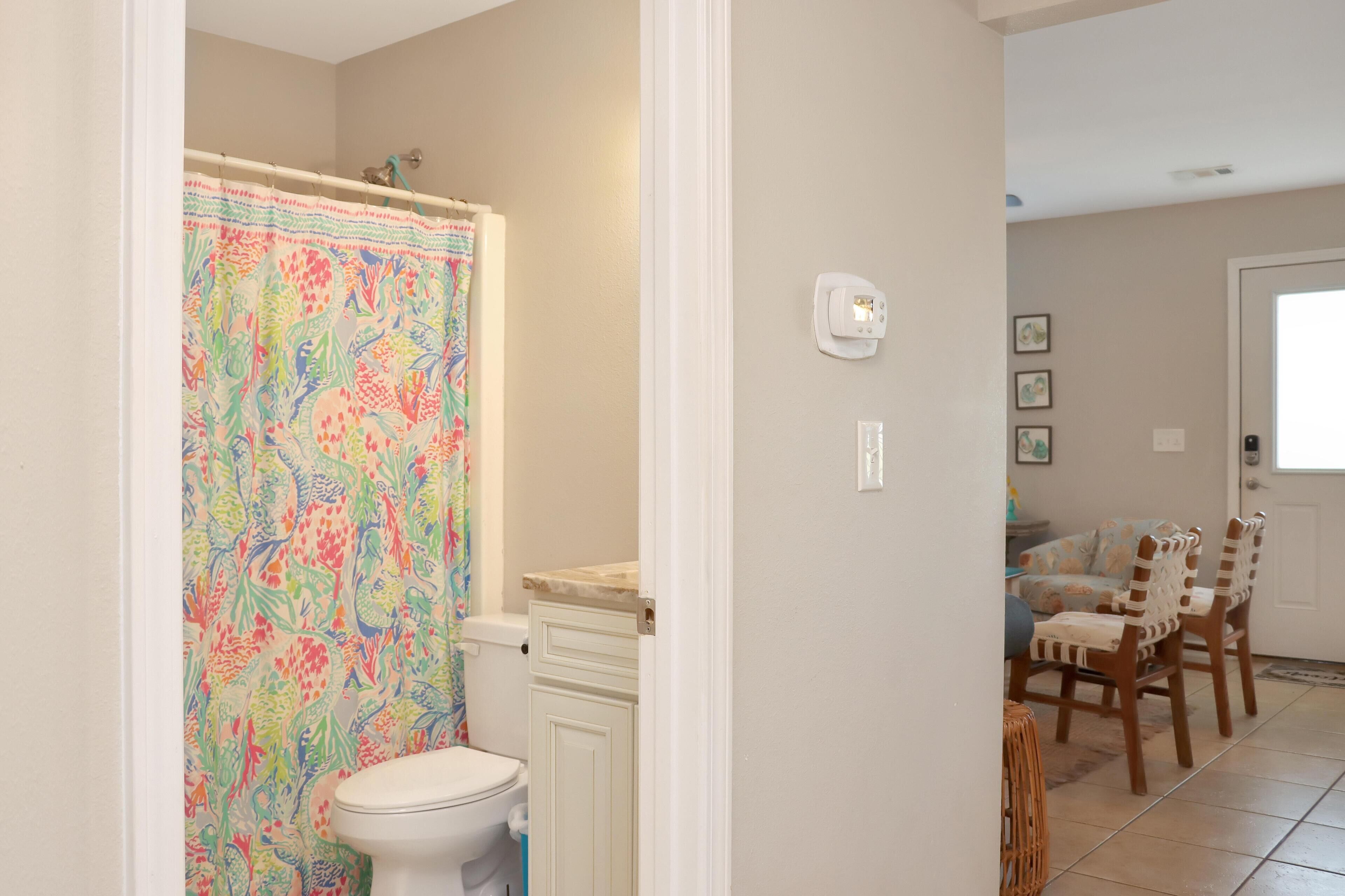 A bathroom with a colorful shower curtain, a toilet, and a cabinet. There is a door leading to a dining area with chairs and a table.