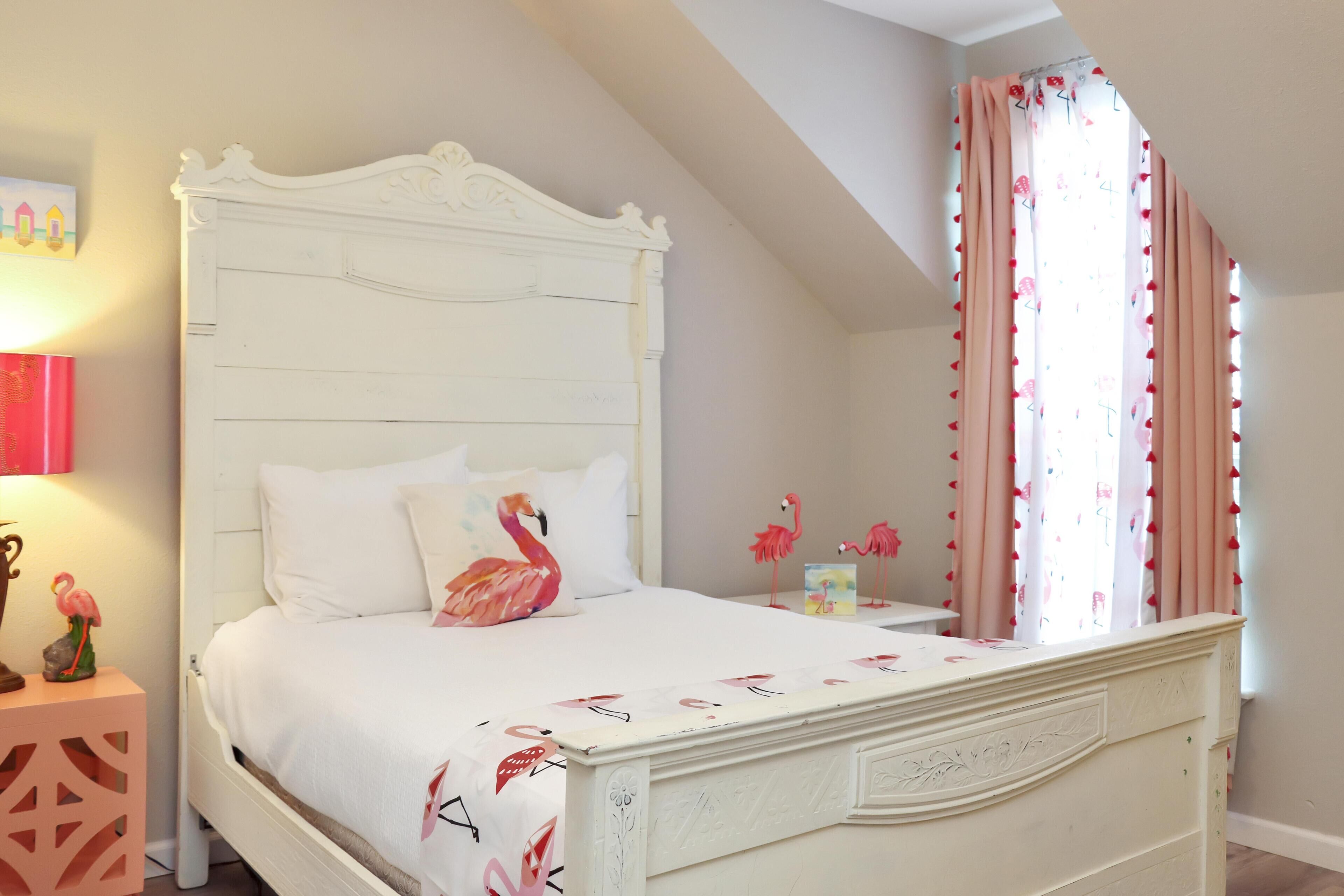 Une chambre avec un lit blanc, une table de chevet, un flamant rose décoratif et une fenêtre avec des rideaux.