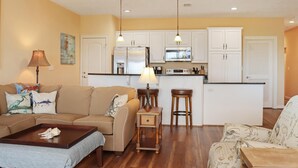 A living room with a sofa, coffee table, and a lamp. There is a kitchen area with white cabinets, a refrigerator, and a microwave. - Club at Cape San Blas 1C - Sandpiper (Port St. Joe)