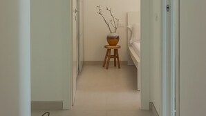 Un couloir mène à une chambre où se trouvent un lit, un petit tabouret surmonté d’un vase et une table en bois avec un panier.