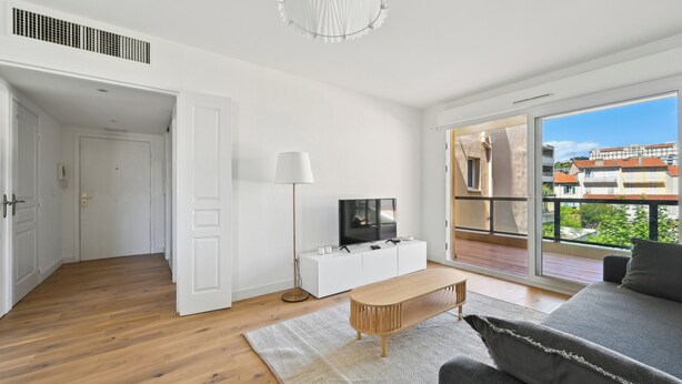 A modern living room with a wooden floor, a white sofa, a TV on a white cabinet, and a large window with a view of buildings. - Appartement Ensoleillé Avec Terrasse à Cannes (Cannes)