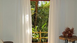 Una habitación con vistas a un jardín a través de una ventana con cortinas blancas. Hay una mesa de madera con un jarrón de plantas secas.