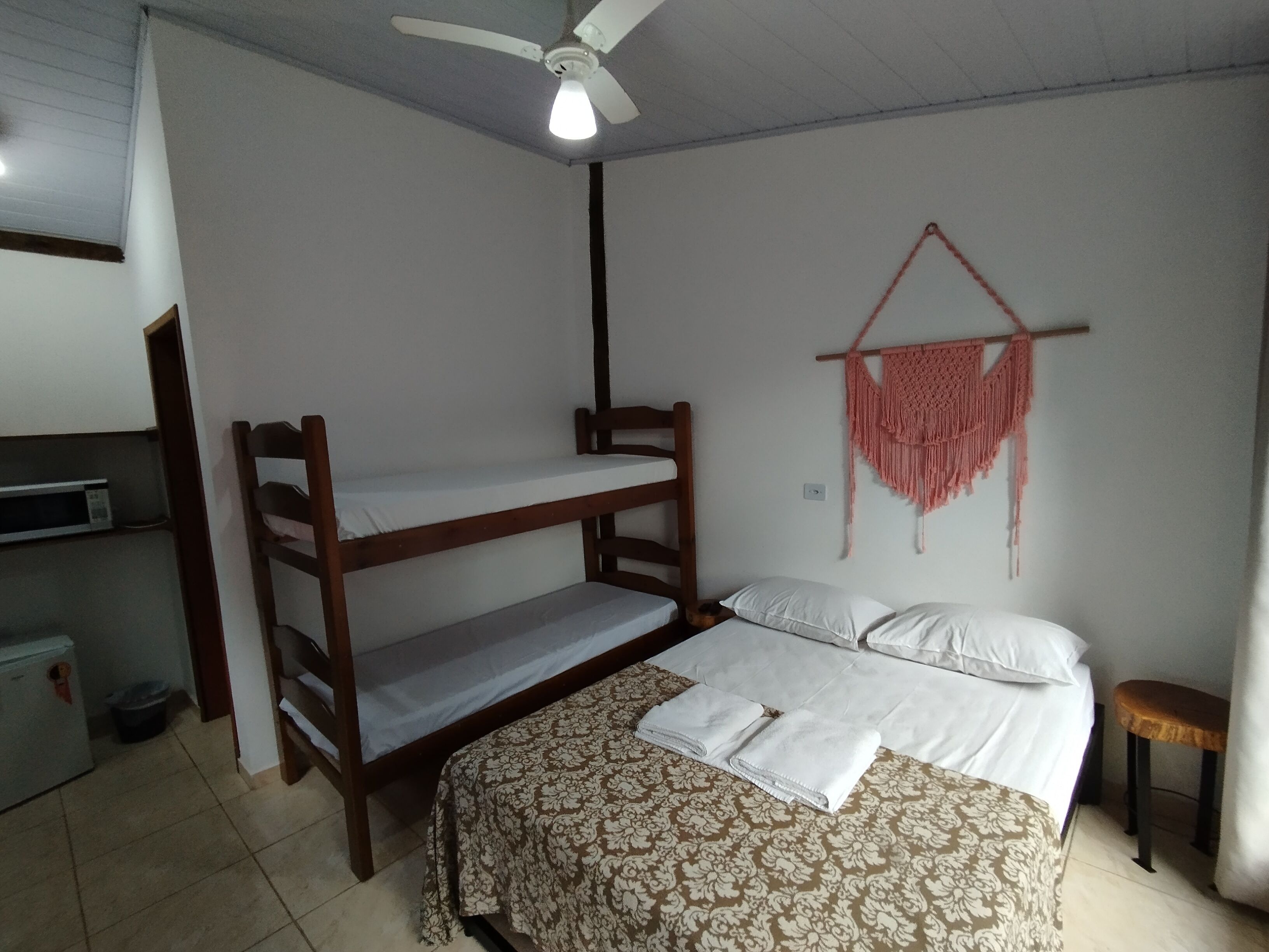 A room with a bunk bed, a desk with a microwave, a ceiling fan, and a macrame wall hanging.