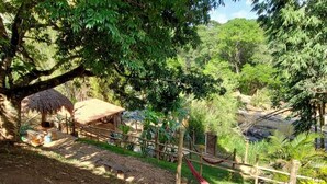 Una hamaca bajo un árbol, una estructura con techo de paja y un jardín cercado.