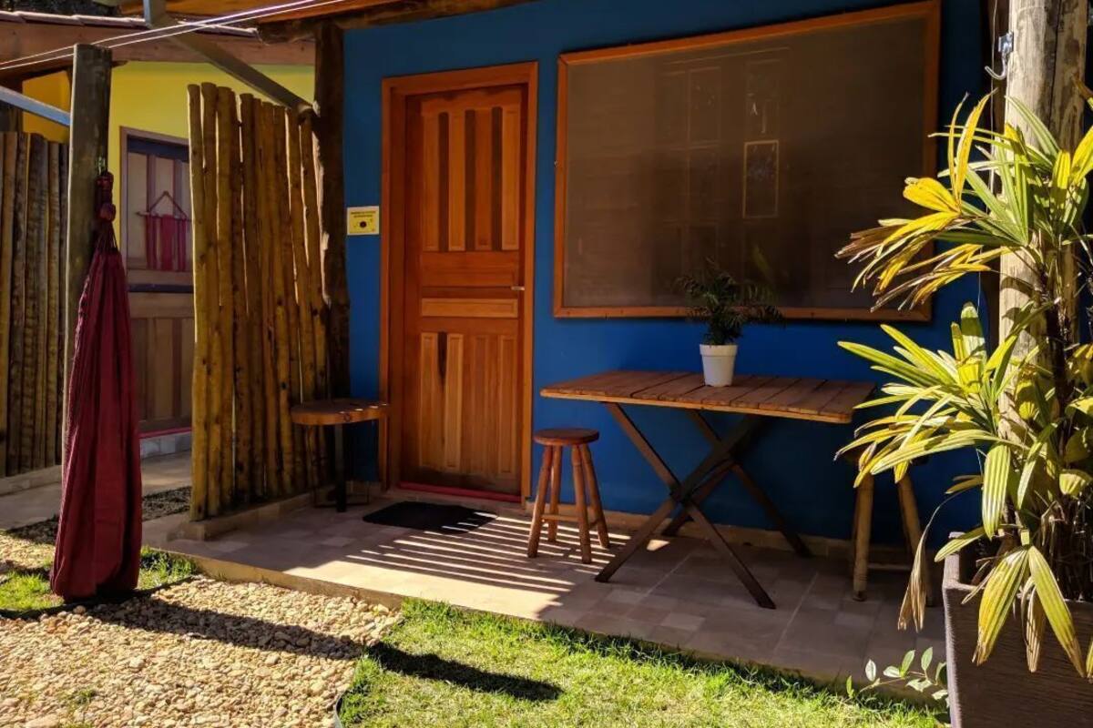 A small, rustic building with a wooden door and window, a small table with a potted plant, and a red garment hanging on a wooden fence.