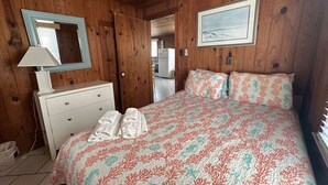Une chambre avec des murs en bois, un lit recouvert d’une literie à motifs floraux, une commode blanche et un ventilateur de plafond.