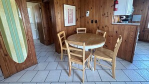 Un intérieur en bois avec une table ronde et des chaises en bois, une planche de surf appuyée contre le mur et un coin cuisine avec des placards et un extincteur.