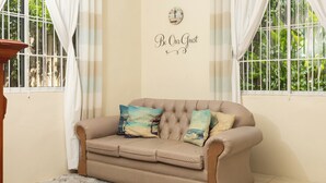 A beige sofa with decorative pillows, a fluffy rug, and a wall with a clock and a sign saying "Be Our Guest". - Adorable 1Bed Cottage Walk to the Beach (St. Ann's Bay)