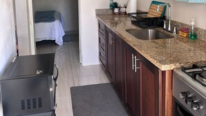 A compact kitchen with dark wooden cabinets, a granite countertop, a sink, and a stove. There are kitchen rules on the wall, a small plant, and a bed in the adjacent room. - Adorable 1Bed Cottage Walk to the Beach (St. Ann's Bay)
