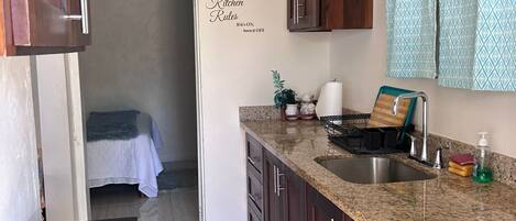 Una cocina compacta con armarios de madera oscura, encimera de granito, fregadero y estufa. Se observan normas de la cocina en la pared, una planta pequeña y una cama en la habitación contigua.